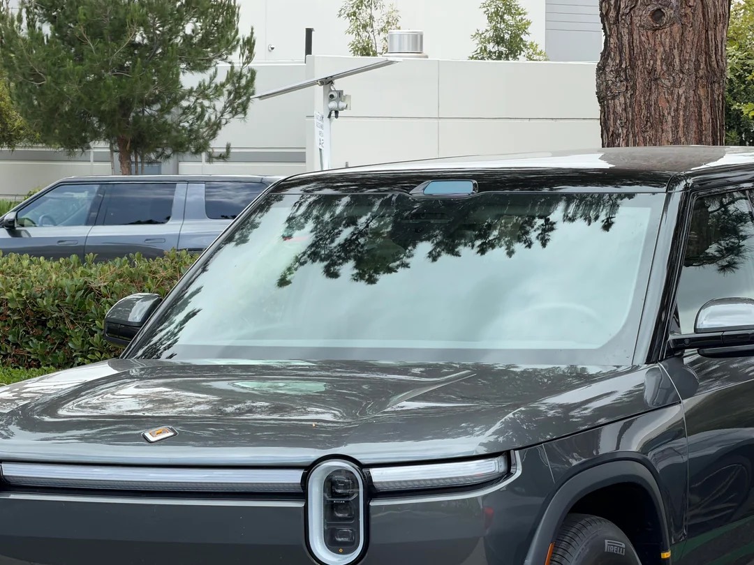 Front half of a dark gray electric SUV parked in a parking lot, with tree reflections on the windshield and a blue SUV in the background.